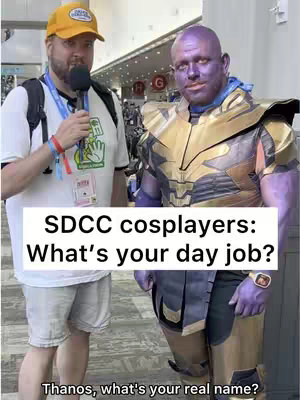 Two men in costume at a comic convention. On the left is a man wearing clothing with a media badge, holding a selfie stick with a microphone and speaking into it. On the right is a man cosplaying as Thanos, a purple-skinned character with golden armor.