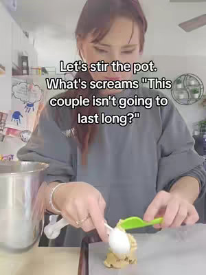 A woman in a gray sweatshirt is shown in a kitchen setting, focusing on her hands as she uses a spoon and green spatula to handle cookie dough on a sheet of parchment paper. Text overlays are added to the picture at the top, suggesting a relationship theme.