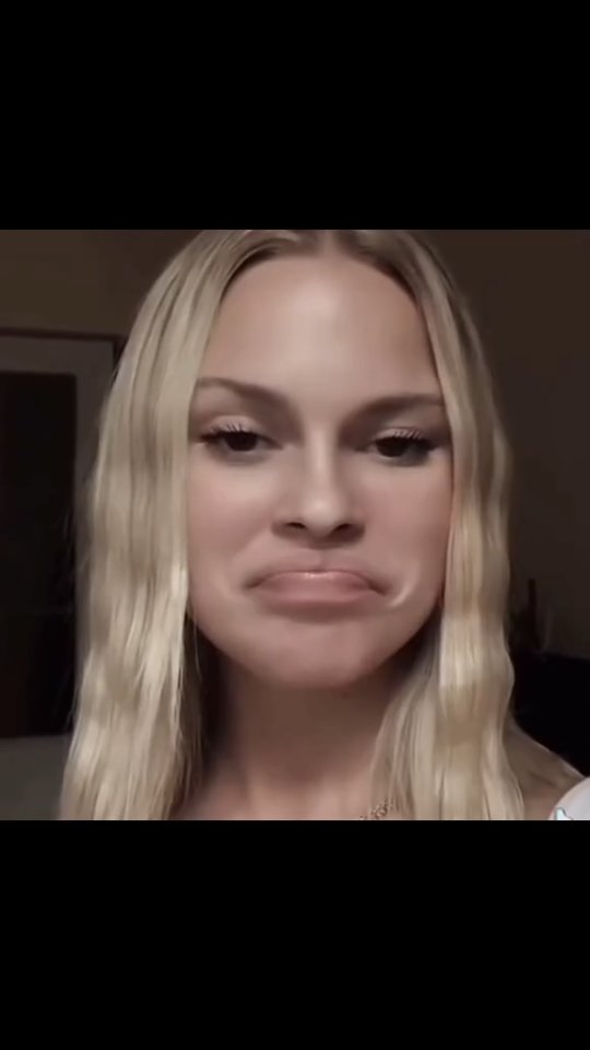 A close-up shot of a woman with long, wavy blonde hair. She has a slightly exaggerated expression, with a subtle downturn of her lips, as if she's playfully making a face. The lighting appears soft, casting a warm glow on the subject, and the background is blurred, drawing focus to the woman's face.