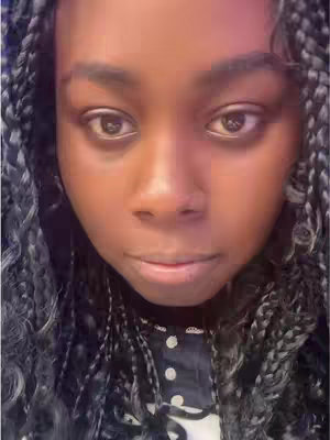 Close-up shot of a woman's face. She has black hair styled in braids, framing her face. Her skin is a warm brown tone. She is looking straight ahead with a neutral expression. The background is dark and indistinct..
