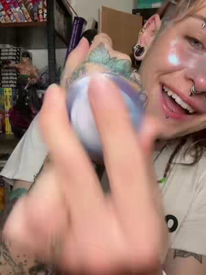 A person with multiple ear piercings, nose piercings and lip piercings is looking at something. The individual wears a white T-shirt and has visible tattoos. The person is holding a blue and white ornament, it appears to be a decorative ball. In the background is a bookshelf with some books and a statue.