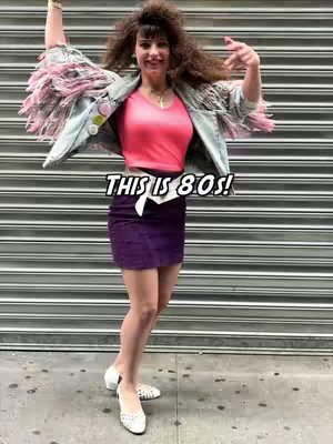 A woman in 80s attire, wearing a denim jacket with pink fringe, a pink top, and a purple skirt, is posing with one arm raised and the other gesturing. The background is a metal roll-up door, and a text overlay reads "THIS IS 80S!"