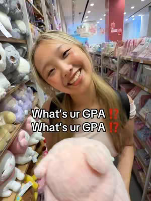 A young woman with long blonde hair is smiling widely, holding a large pink plush animal, likely a stuffed animal, in a brightly lit retail setting. Behind her are shelves filled with merchandise, suggesting a store. The text overlay asks "What's ur GPA ??" repeated.