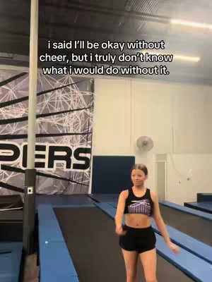 The cover image captures a young woman in a cheerleading gym setting. Positioned centrally, she is wearing a black sports bra and shorts. Behind her, a large banner with the partial word "PIERS" is visible, and equipment like a trampoline and safety padding fill the background, indicating a training environment. The color palette leans toward neutral tones, with the subject and the setting uniformly lit.