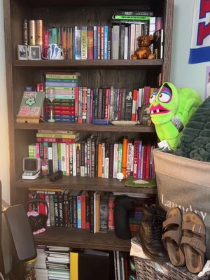 A wooden bookshelf filled with books, decorative items, and personal belongings. The shelves hold a diverse collection of books of varying sizes and colors. There are small objects and pictures on top, and a stuffed animal. A lime green puppet-like creature sits on a basket. Shoes and accessories are in front of the shelf.