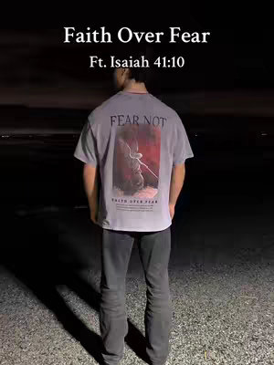 A person is shown from the back, wearing a gray t-shirt with a graphic on the back. The person is standing on a gravelly surface, and the background is dark, suggesting nighttime.  The t-shirt graphic has the words "FEAR NOT" at the top, a center image of a figure with a red color scheme, and it reads "Faith Over Fear" at the bottom.  Above the person, the hooks "Faith Over Fear Ft. Isaiah 41:10". The lighting is muted, with the focus on the person and the message on the shirt.