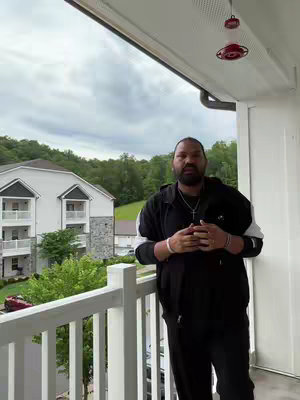 A man in a black hoodie stands on a balcony with his arms crossed and a pendant cross necklace around his neck. A white railing and a building are in the background.