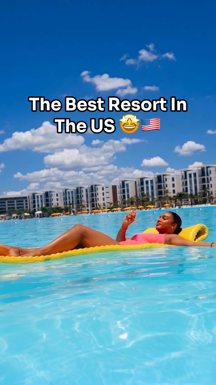 A woman lies on a yellow floating raft in a pool of blue water at a resort, with a backdrop of clear blue sky with white clouds and a row of hotel buildings. Text overlay indicates the resort is in the US.