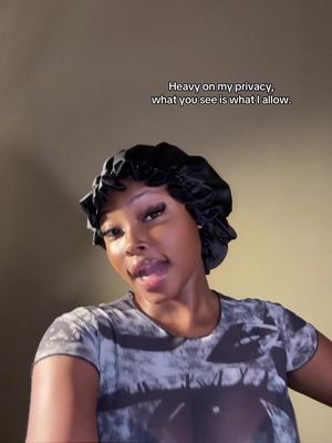 A woman with dark skin is positioned in the center of the frame, facing the camera. She's wearing a black satin head covering, a patterned t-shirt. The background is out of focus. Text overlay at the top reads, \"Heavy on my privacy, what you see is what I allow.\"