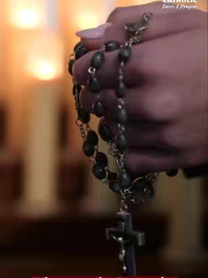 A hand holding a rosary, with the crucifix at the bottom. Behind the hand are blurred images of candles and a soft, warm light.