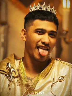 A man with dark hair and a crown on his head sticks out his tongue. He is wearing a gold and white outfit. He has a playful expression. The background is a blurred, warm-toned interior.