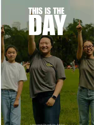 The cover image depicts three young women standing on a grassy field, with a lush green background and a clear sky. The central figure is smiling and pointing upwards with one finger, and the other two women are also raising their arms. Large white text that reads, "THIS IS THE DAY" is superimposed at the top of the image, with a church logo beneath. The overall composition is bright and celebratory.