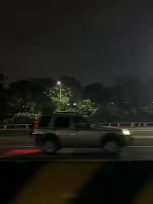 A nighttime shot of a road with a silver SUV driving on the right side. The scene is dark, with streetlights illuminating the trees in the distance. There is a guardrail on the left, and the image is slightly blurred, suggesting motion.