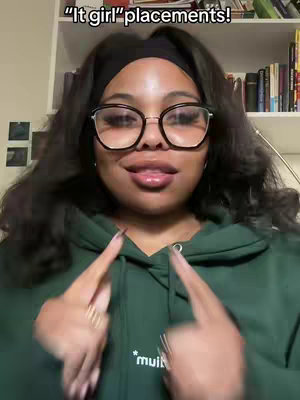 A woman with dark hair, wearing glasses and a green hoodie, is pointing with both index fingers towards her face. The background includes a bookshelf.
