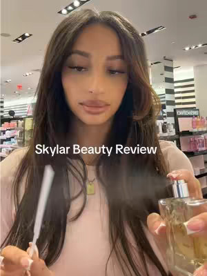 A woman with long dark hair is centered in the frame, holding a perfume bottle and a white stick, likely a testing strip or applicator. The background appears to be a retail store, possibly a beauty or fragrance outlet, with shelving visible. The woman appears to be spraying the perfume.