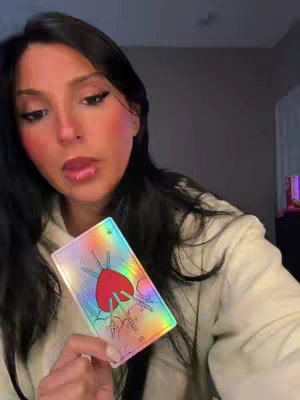A young woman with long dark hair is holding a tarot card. She's wearing a light-colored hoodie. The background is dark, and the card is iridescent with a depiction of a heart and swords.