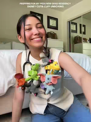 A young woman wearing a white top and blue jeans sits indoors, smiling at the camera. She has a stethoscope around her neck and a collection of small stuffed animals hanging from it. The background features a white couch, a mirror, and a dresser.