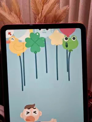 A tablet screen displays a colorful mobile game scene.  Above are cartoon shapes: a yellow duck-shaped balloon with a long blue string; a green four-leaf clover balloon; a yellow star balloon; and a green frog balloon. Underneath is a cartoon character with a surprised expression.