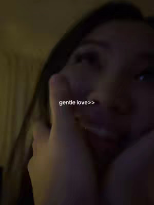 A close-up shot of a person, likely a young woman, smiling with her head resting on her hands. The lighting is dim, creating shadows and a sense of intimacy. The background is dark and out of focus, with a warm, yellowish hue. The focus is on the person's face, with soft, warm lighting on the skin. The text overlay reads 'gentle love>>'.