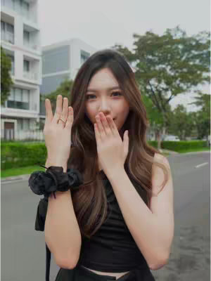A young woman stands in front of a residential street with manicured medians and buildings in the background. She is wearing a black top and holding her hands up towards her face. Her expression suggests a playful gesture.