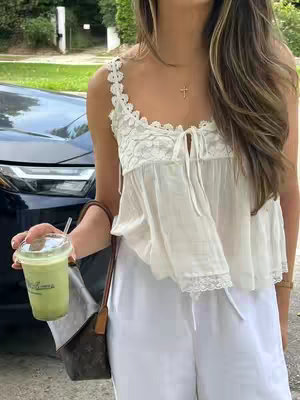 A young woman is captured in a selfie-style shot. She is dressed in a white, lacy top and matching wide-leg pants, holding a green drink in a clear plastic cup with a straw. A stylish brown purse is slung on her shoulder, and she wears a delicate necklace. She is standing near a parked black car, with a lush green background and sidewalk. The lighting is natural, soft, and creates a warm atmosphere.