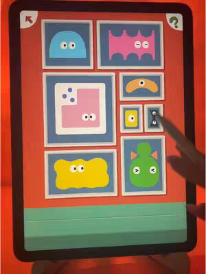The image shows a tablet displaying a puzzle game. The game features different colorful shapes and animated forms within picture frames. The tablet has a black border and rests against a light brown surface against a red backdrop. A finger is pointing to a frame of the game.