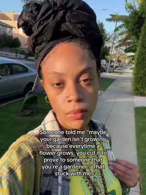 A selfie-style shot featuring a person of color standing outdoors, with houses and greenery in the background. The person is wearing a head wrap, a plaid shirt, and a t-shirt, and has jewelry, including hoop earrings and a nose ring. Text overlay is placed at the bottom of the screen.