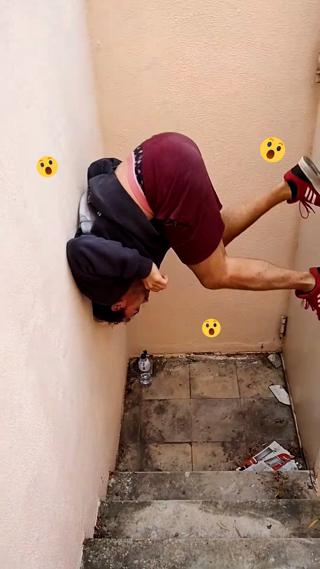 A man is upside down, leaning against a light pink wall with his head pressed against it, his legs are up toward the right on the frame. He is wearing a black hoodie, burgundy shorts, and red Adidas sneakers. The wall shows three yellow emojis each with a surprised expression. In the background, there are stairs and a water bottle.