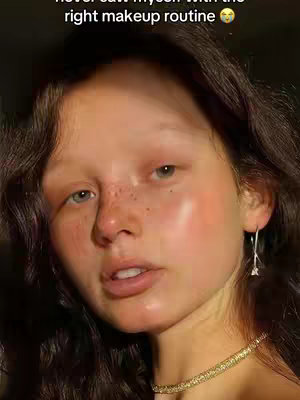 The cover image is a close-up selfie. The focus is on a person's face, which has dewy-looking skin, freckles, and a soft glam makeup look featuring blush and luminous highlights. The person has dark wavy hair, and is wearing a delicate gold necklace and a single silver earring. The background is dark, and lighting is warm and natural.