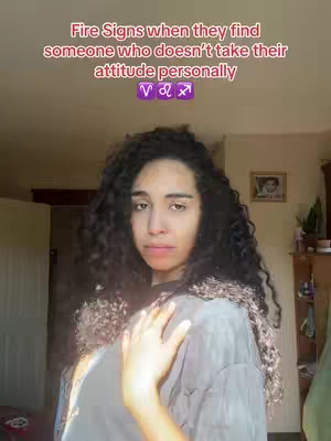 A medium shot features a woman with curly dark hair, positioned slightly off-center against a blurry indoor backdrop. She wears a grey top and looks towards the camera. Above her, text overlays in a bold, red font that reads: "Fire Signs when they find someone who doesn't take their attitude personally". Below that, the symbols for Aries, Leo and Sagittarius are presented in a purple box.