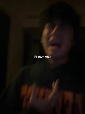 A blurry, underexposed image featuring a young man, captured in a low-light setting. The man is wearing a dark-colored top and a black cap, with his face partially visible and out of focus. A text overlay displays the phrase "i’ll love you" in white font. The background is obscured, suggesting an indoor environment. The man appears to be speaking or singing, with his mouth open. The overall image has a dark, emotional tone.
