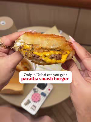 Close-up shot of a smash burger held in hand, with a paratha bun, cheese dripping and onions. A phone with a pink design is visible in the background.