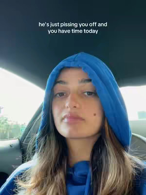 A woman with long brown hair is seated in a car wearing a blue hoodie with the hood up. Text overlayed on the image reads "he's just pissing you off and you have time today". The background is blurred and includes hints of other cars and possibly a city atmosphere.