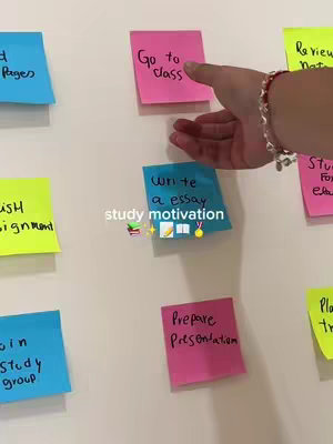 A person's hand reaches towards a pink sticky note that says "Go to class". Several other sticky notes are stuck on the wall. Some say "Write a essay" and "Prepare Presentation". The notes have handwriting. The background is a white wall.