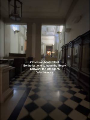 An interior shot of a long hallway in a building. The floor is a black and white checkered pattern. On either side are pillars or large walls with rectangular openings. There are text overlays on the image in white font.