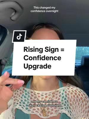 A woman is inside of a car sitting in the driver's seat, looking toward the camera. The background is out of focus. Large bold white text covers the top half of her face: "Rising Sign = Confidence Upgrade." At the top left corner of the screen is a TikTok logo. Below the text, the woman's chest is visible, showing a light blue tank top and a patterned, off-white top over it. Text along the bottom of the screen reads, "and my confidence skyrocketed overnight."