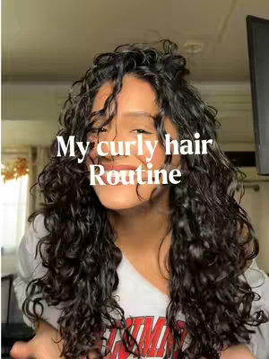 A selfie-style photo featuring a person with dark, long, curly hair. The person is smiling and framed from the chest up, with their hair prominently around their face. The background is softly blurred, suggesting an indoor setting. Text overlayed on the image reads 'My curly hair Routine' in white text.