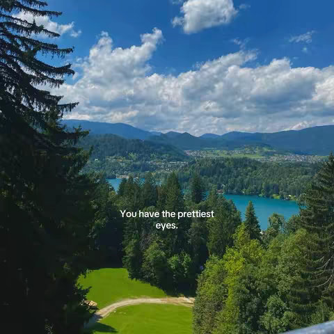 A scenic view of a lake and surrounding mountains under a partly cloudy sky. The foreground includes lush green trees and a grassy area. Text overlay reads, 'You have the prettiest eyes.'