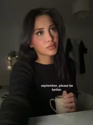 A woman with long dark hair is looking up, seemingly thoughtful. She's holding a mug.  Text overlayed says 'september, please be better.' The lighting is dim.