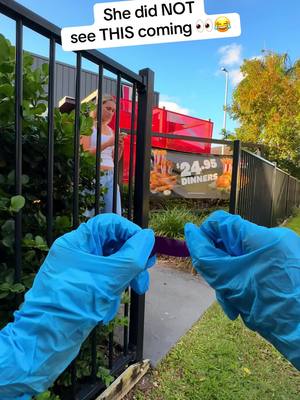 The image shows a first-person view of someone holding a small purple object, likely a pen, near a black metal fence. The person's hands are covered in blue gloves. Behind the fence, a woman is seen. In the background, there's a KFC sign, a building, and greenery under a bright blue sky. Text overlayed reads, 'She did NOT see THIS coming.'