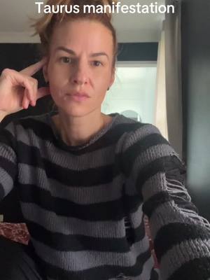 The image features a woman looking directly at the camera, seemingly in thought. The background is soft, with curtains and a wall dimly visible. The woman is wearing a striped sweater, and her hair is pulled back. A text overlay at the top reads "Taurus manifestation". The overall lighting appears natural.