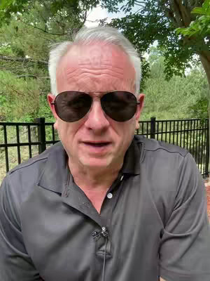 A medium shot of a man wearing sunglasses, posing in front of a blurred background of trees and a fence. The man is wearing a gray shirt, and a small microphone is clipped to his shirt.