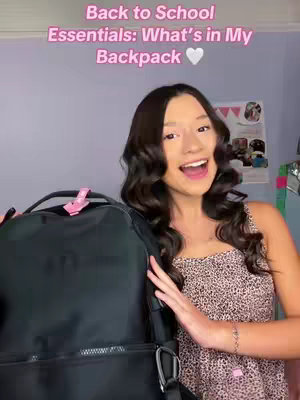 A young woman with long, wavy dark hair smiles widely, holding a black backpack towards the camera. The background is a light purple wall. Text overlay at the top reads 'Back to School Essentials: What's in My Backpack', with a white heart.