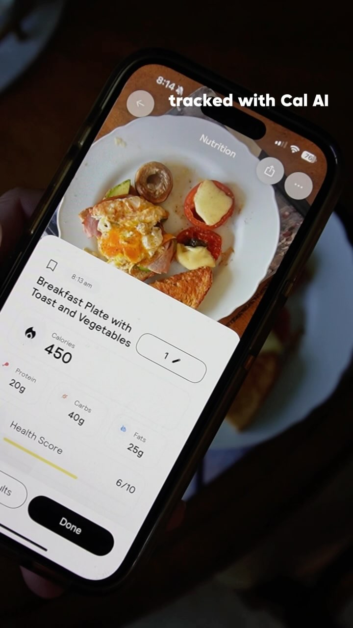 A smartphone displaying a food tracking app with a plate of food, including an egg, toast, and vegetables. The phone is held over the plate, suggesting the app is being used to record the meal. The background is slightly blurred, showing a wooden table, enhancing the focus on the phone's screen.