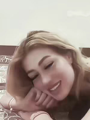 A close-up shot of a fair-skinned woman lying down, propped up on her elbow, looking towards the camera with a slight smile. Her blonde hair is tousled around her face, and her expression suggests a sense of comfort and ease. The background is blurred, suggesting an indoor setting.