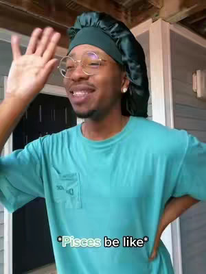 A person with brown skin stands outdoors with one arm raised and a smile. They wear a teal t-shirt and a green satin head covering, and round glasses. The background shows a white house.