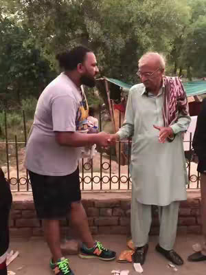 Two men shaking hands amidst a backdrop of greenery and rustic structures. The man on the left is dressed in casual attire, including a grey t-shirt, dark shorts and sneakers. The man on the right, who appears older, is in a traditional light green outfit, and carries a patterned scarf on his shoulder.