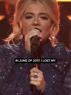 The image features a fair-skinned woman with blonde hair, likely in a performance setting. She's holding a microphone close to her mouth, singing or speaking. The background is dark, likely a stage, with hints of warm lighting. Text is overlaid at the bottom: "IN JUNE OF 2017, I LOST MY". The women wears a navy blue sequined top and large earrings.