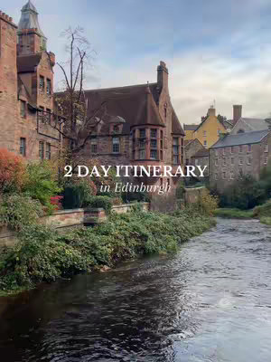 An eye-level shot of a serene river scene with various buildings lining the water's edge under an overcast sky. The foreground features the dark, reflective surface of the river. On the left, a tall, brown stone building and other buildings are visible, with lush green shrubbery. The text overlay reads "2 DAY ITINERARY in Edinburgh".