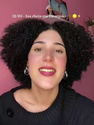 A woman with curly black hair and hoop earrings is smiling at the camera. The background is a solid pink. Text overlay reads: '12/05 - lua cheia em escorpião' with a sun and lightning bolt emoji.
